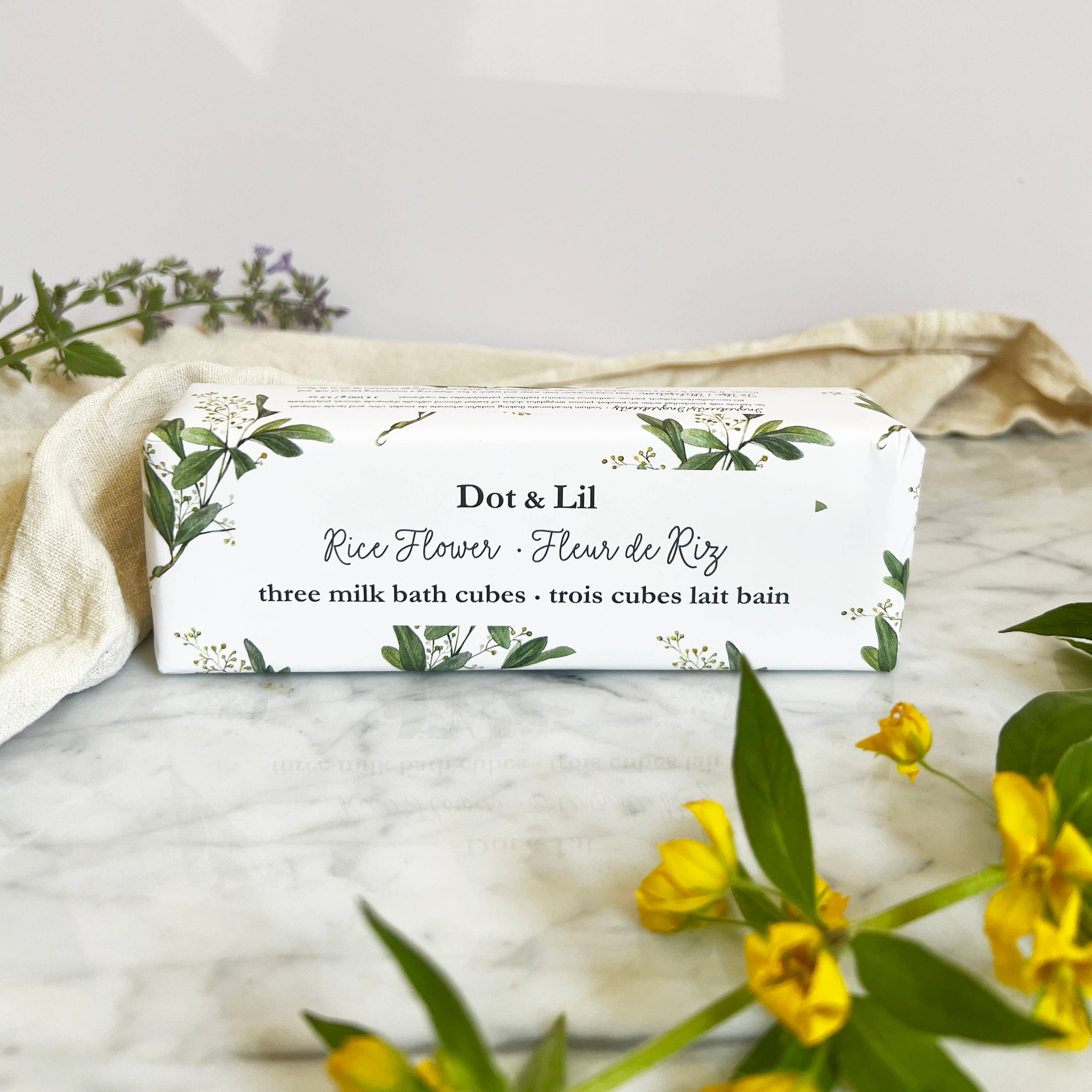 Box of Dot & Lil rice flower milk bath cubes on a marble surface with flowers and leaves.