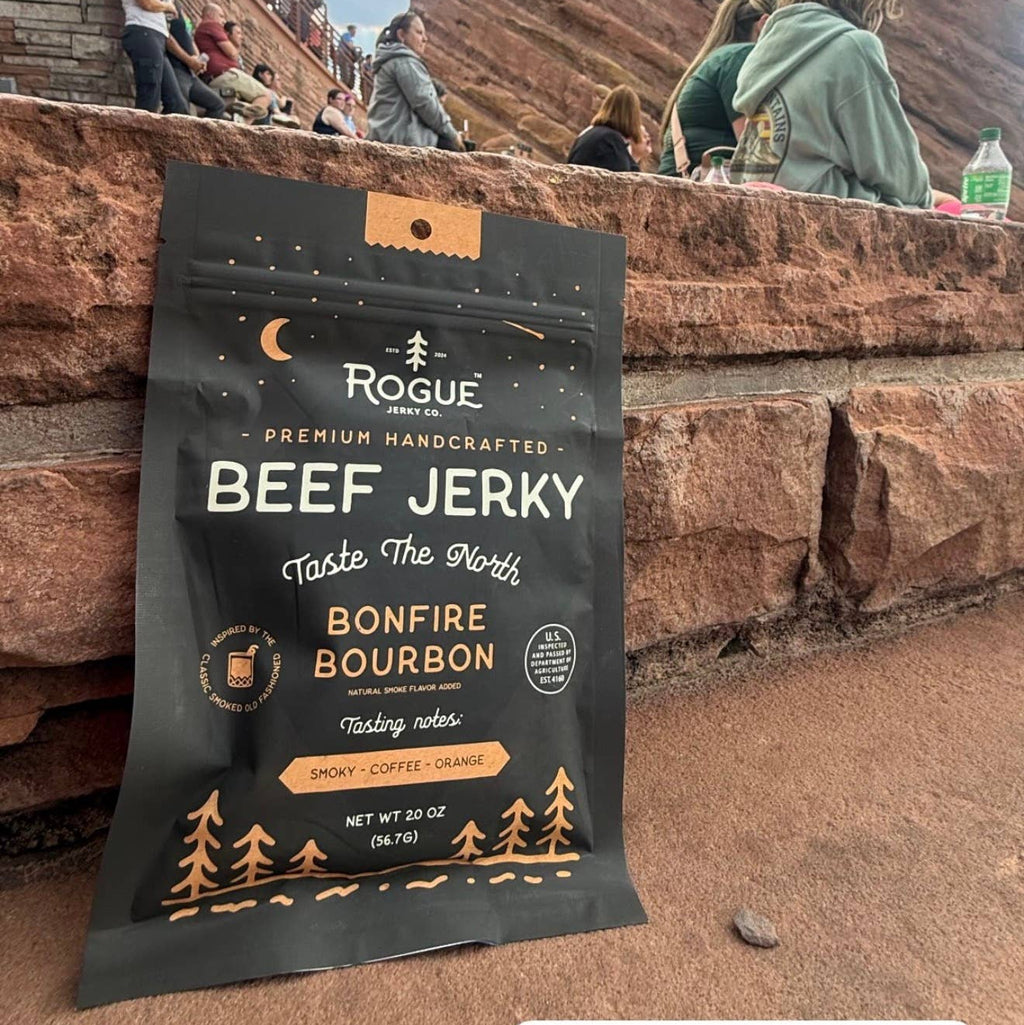 A bag of Rogue Beef Jerky Bonfire Bourbon is displayed in the foreground at Red Rocks Park and Amphitheatre, with the red rock canyon walls and people sitting in the background under a blue sky.
