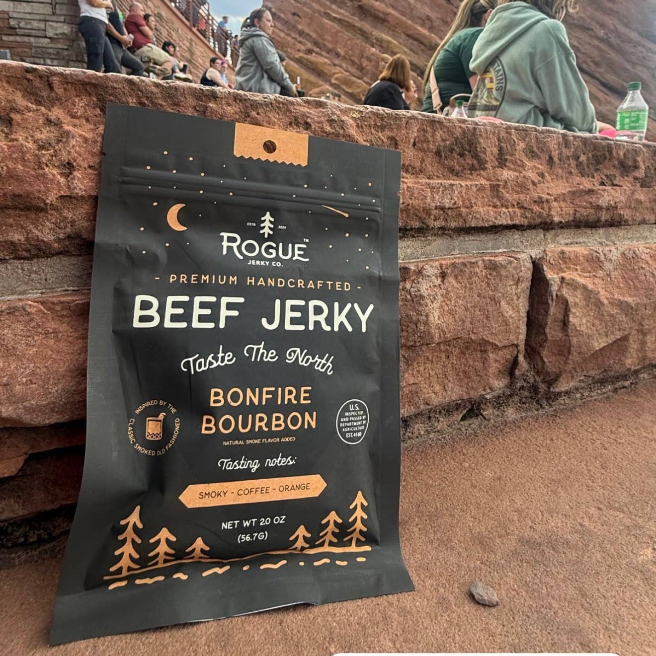 A bag of Rogue Beef Jerky Bonfire Bourbon is displayed in the foreground at Red Rocks Park and Amphitheatre, with the red rock canyon walls and people sitting in the background under a blue sky.