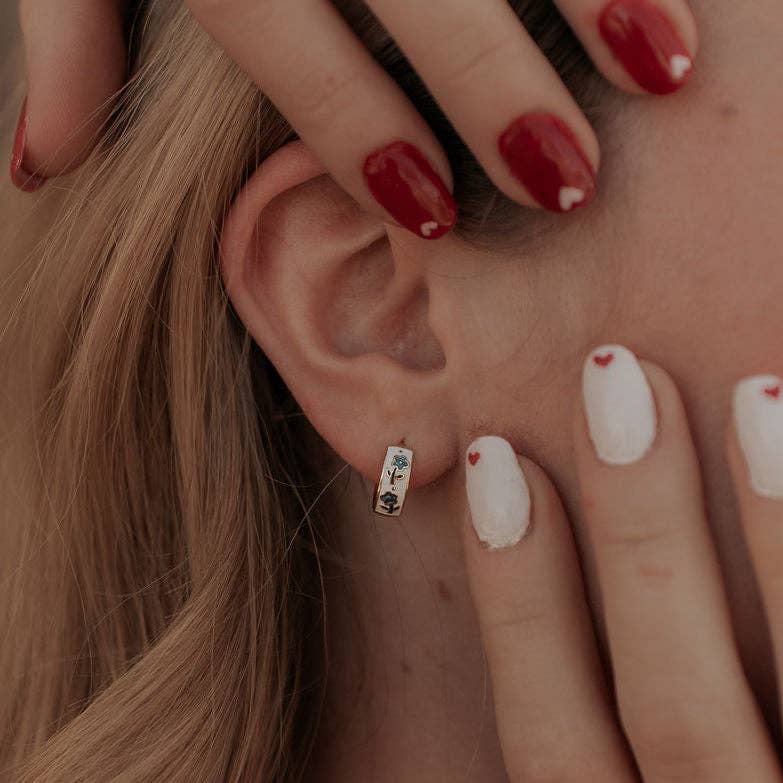 Close-up of an ear with a small earring, hand with red nail polish touching the ear, and heart designs on nails.