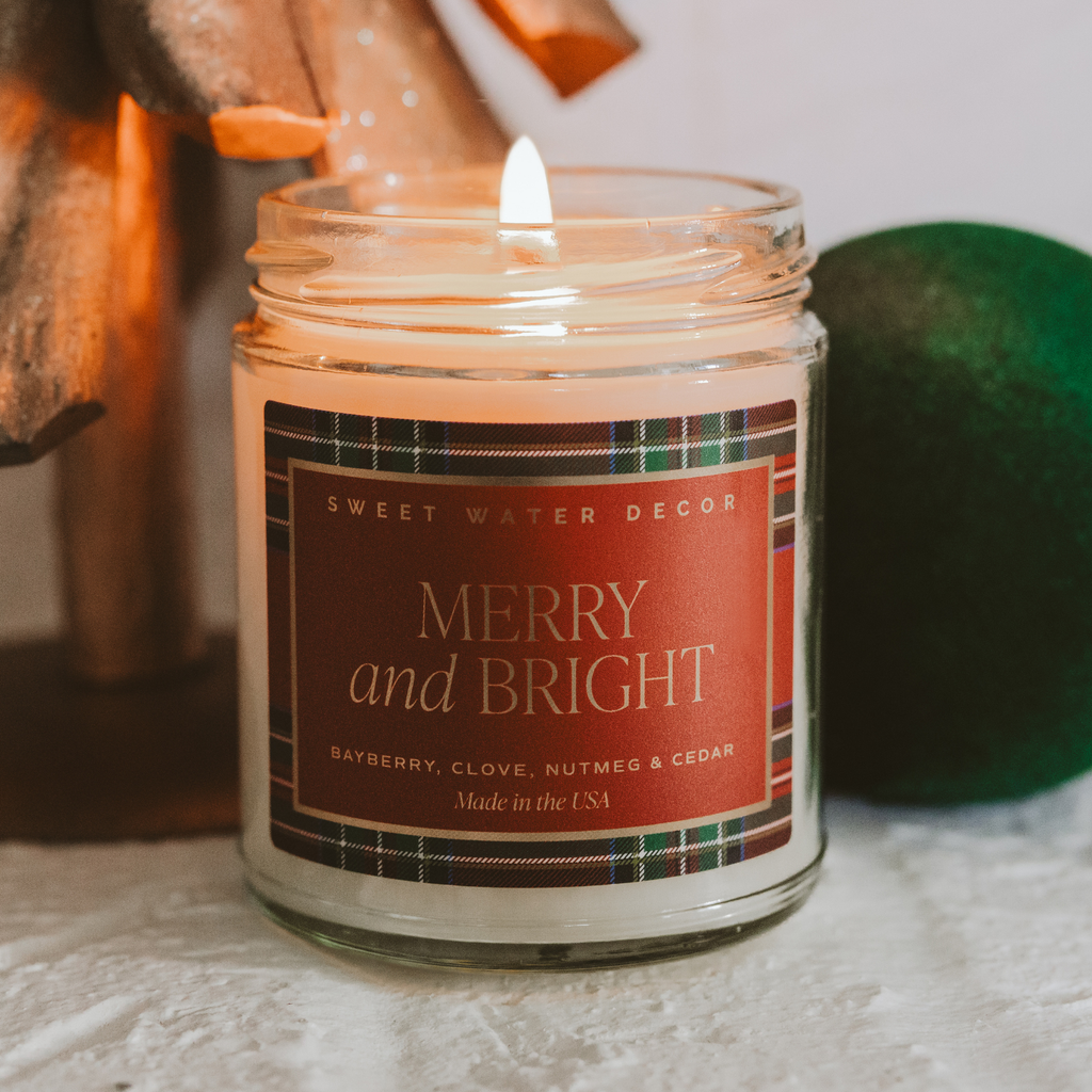 Lit Merry & Bright Christmas candle in a clear glass jar with a red and green plaid label beside holiday decoration