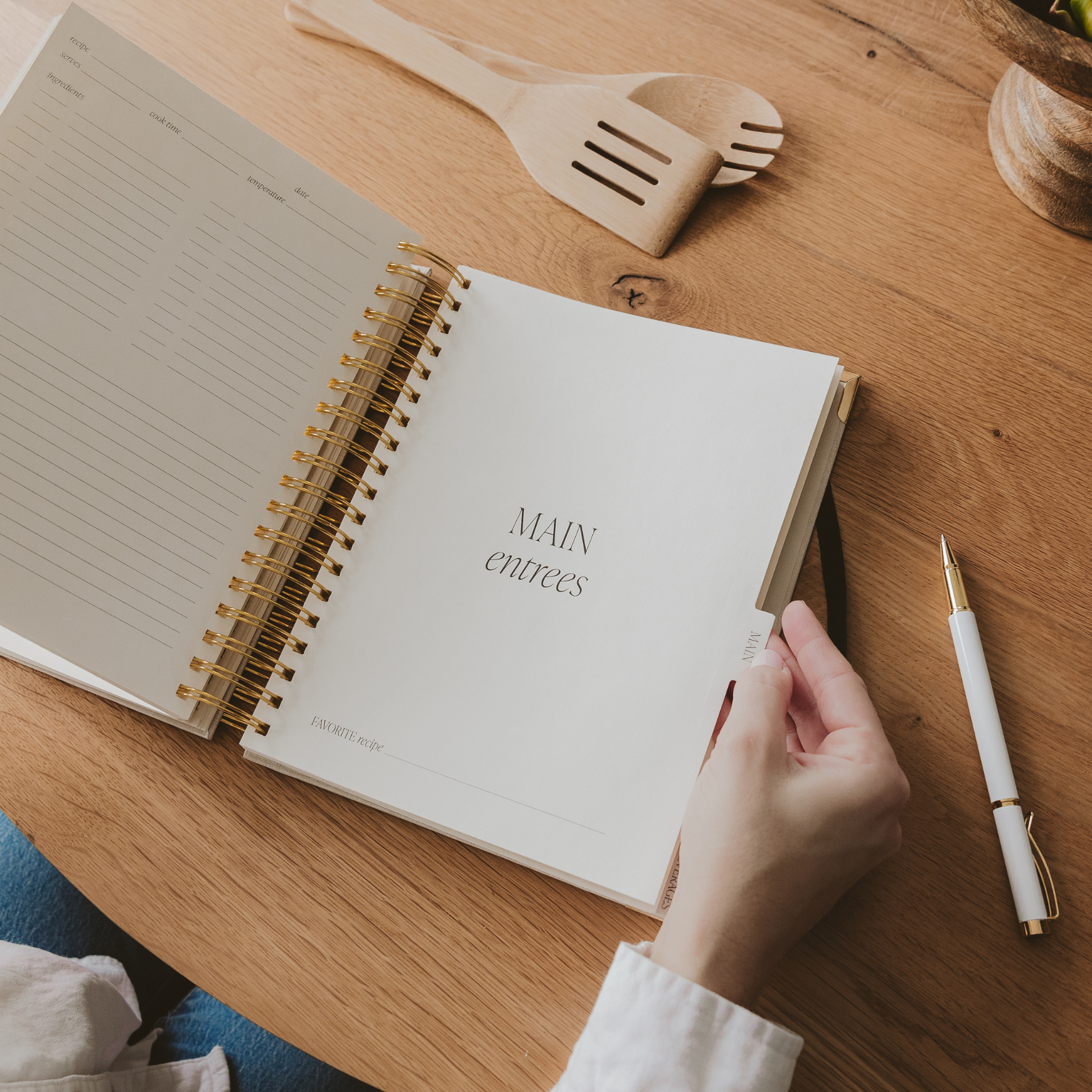 Open notebook on a wooden table with a hand holding a pen, wooden spatula, and small container in the background.
