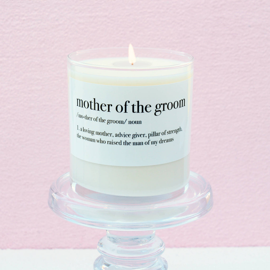 Mother of the Groom Definition Soy Candle in a white glass jar with a meaningful definition-inspired label, displayed on a clear pedestal against a soft pink background.