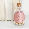 Glass bottle with cork lid containing pink matches and a decorative label on a white surface.