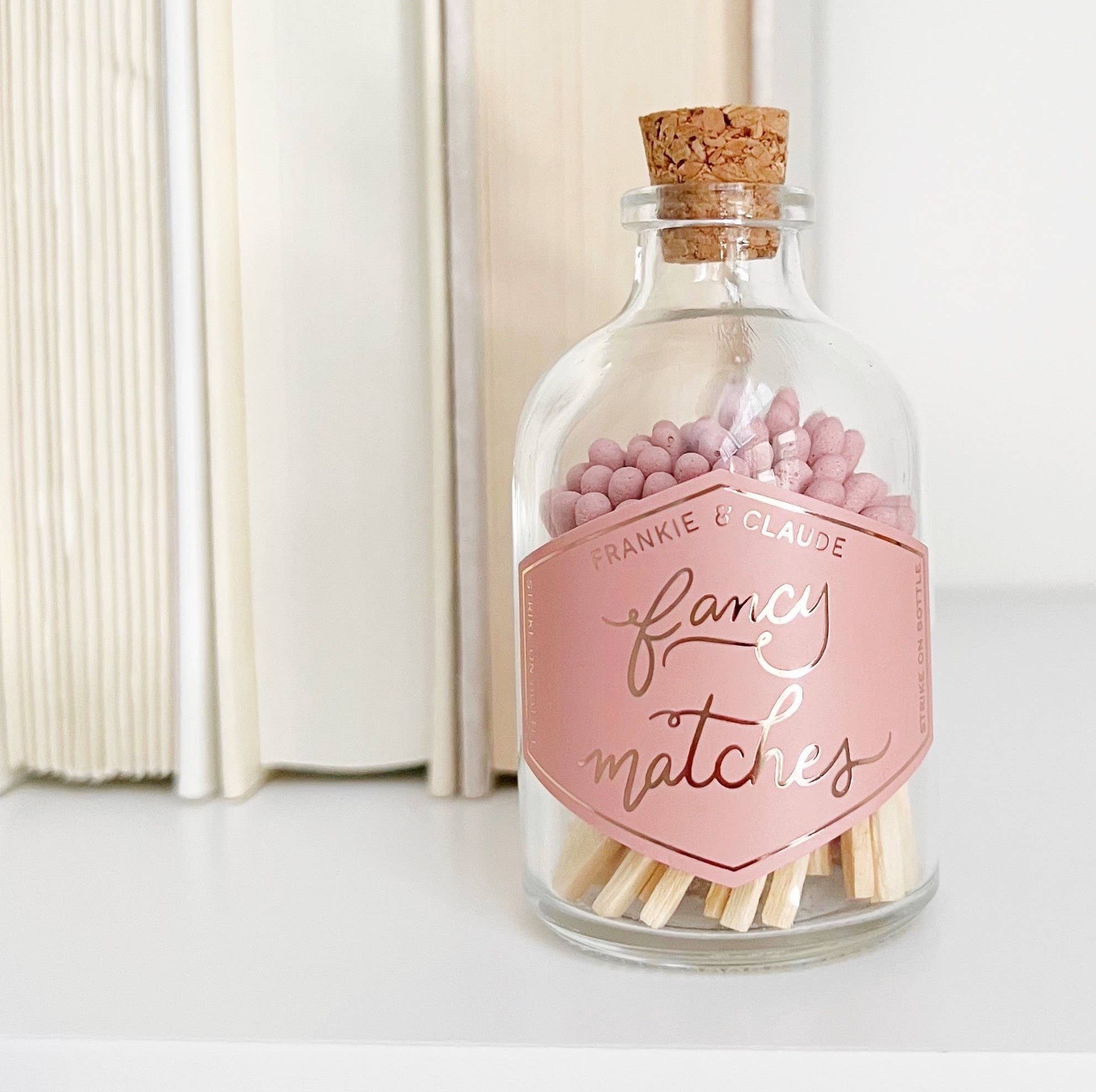 Glass bottle with cork lid containing pink matches and a decorative label on a white surface.