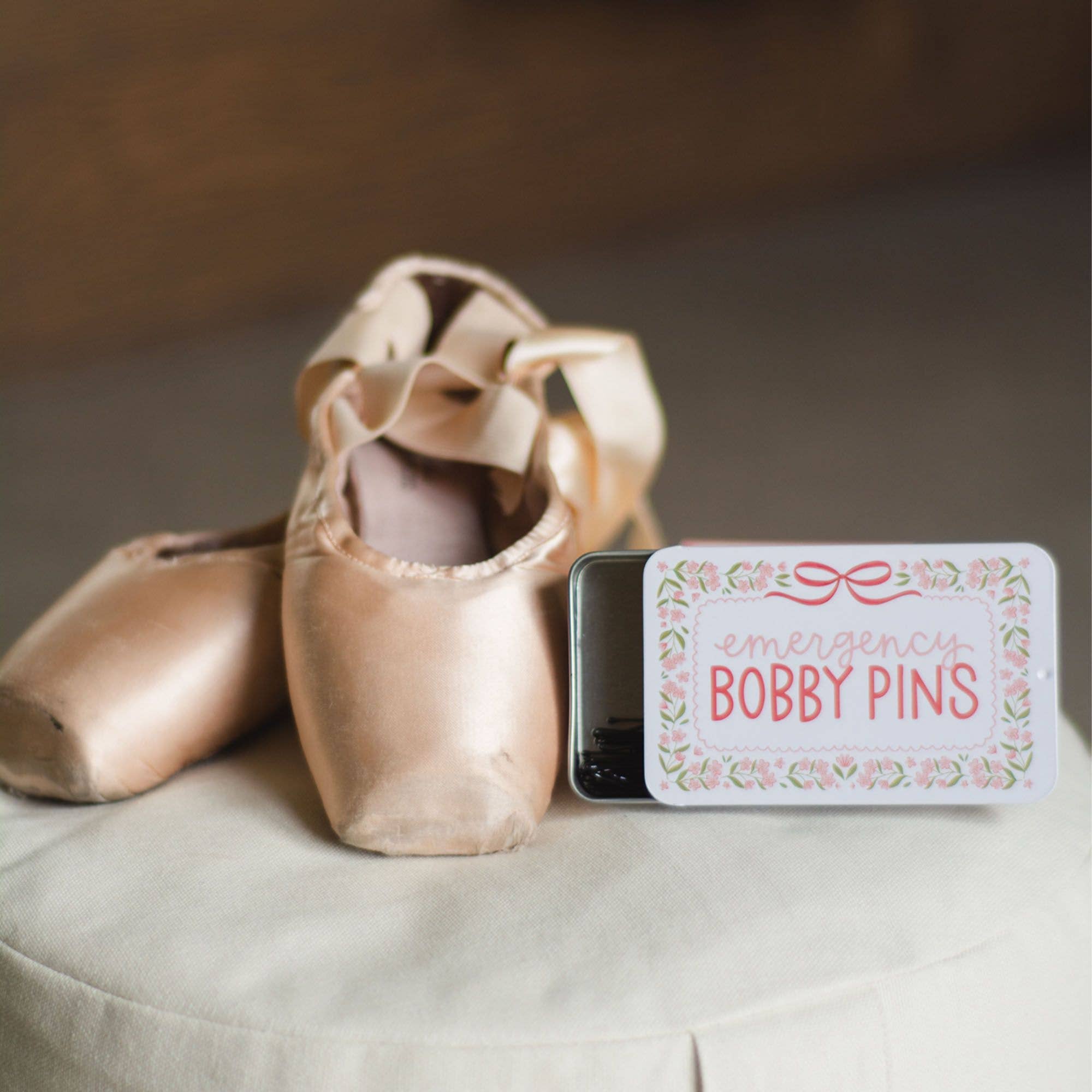 Pair of ballet shoes with a container labeled 'Emergency Bobby Pins' on a neutral background