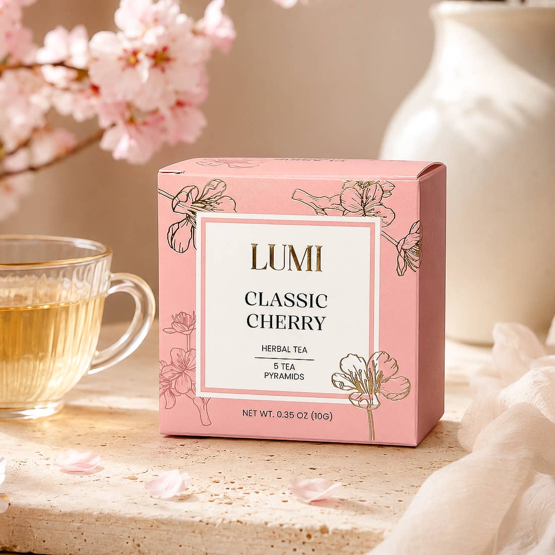 A pink box of LUMI Classic Cherry herbal tea sits on a stone surface beside a glass cup of tea, with soft pink blossoms and a white vase in the background.