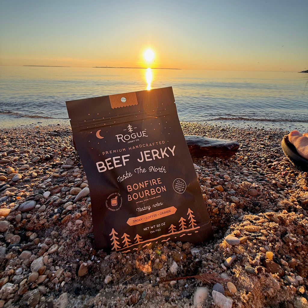 A bag of Rogue Handcrafted Beef Jerky, Bonfire Bourbon flavor, standing upright on a pebble beach at sunset with the ocean in the background.