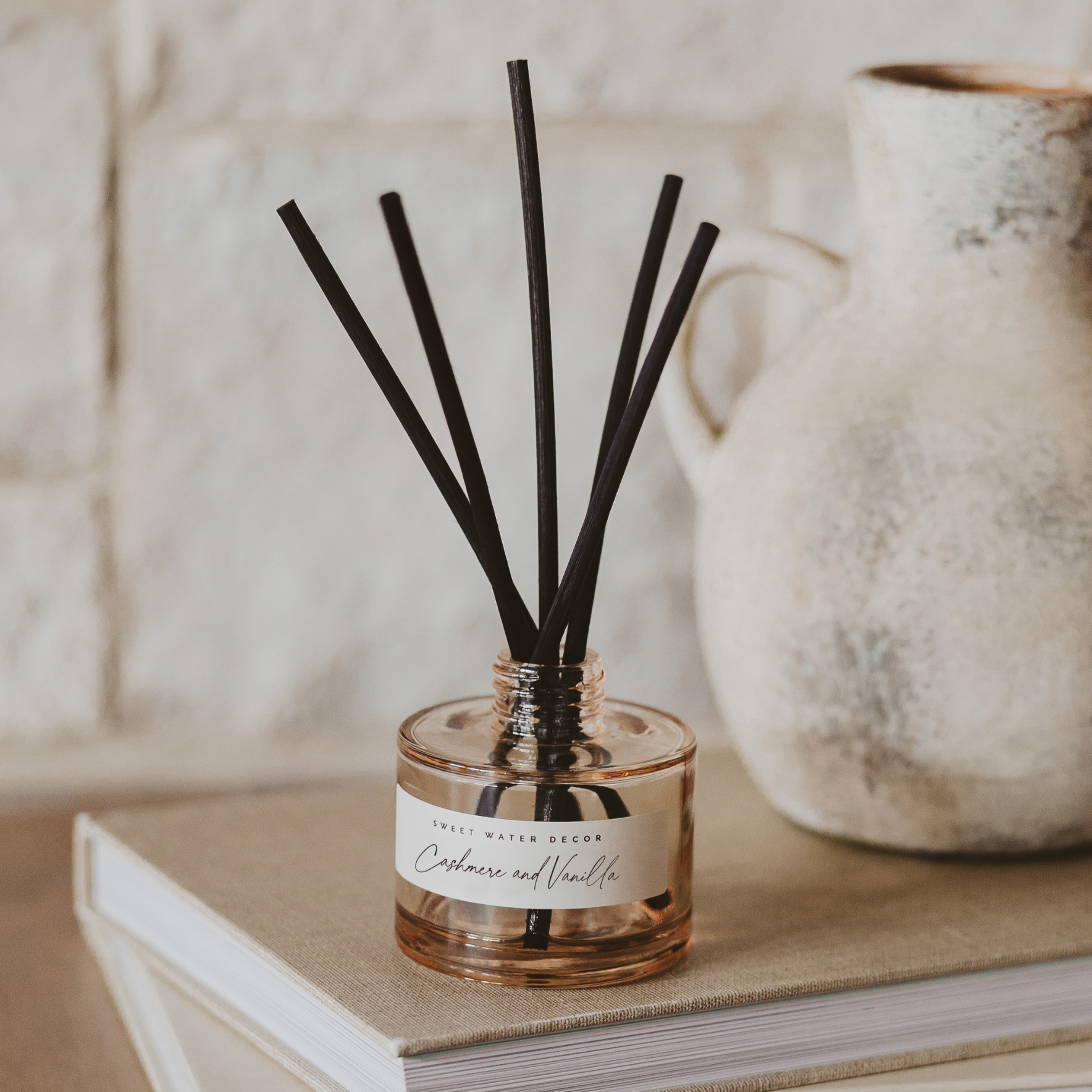 Reed diffuser with black sticks in a clear container on a book with a vase in the background