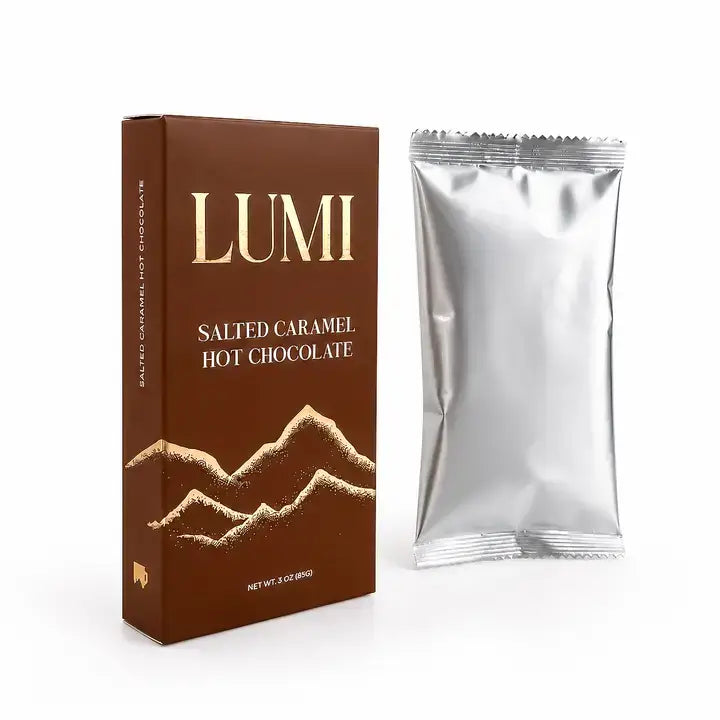 Box and silver pouch: “Angled view of LUMI salted caramel hot chocolate brown and gold 3 oz box beside a sealed silver pouch.