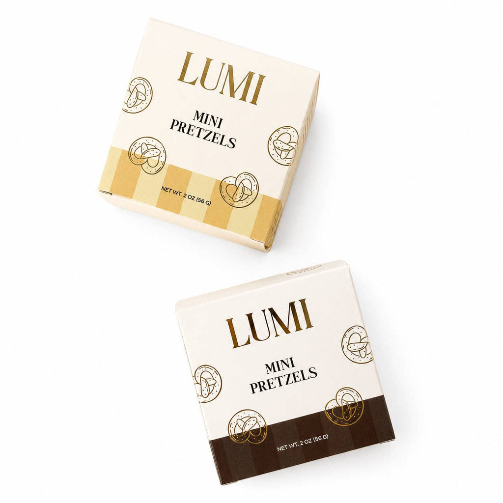 Two LUMI Mini Pretzels 2 oz boxes with gold lettering, one with a yellow striped band and one with a dark brown band, on white.
