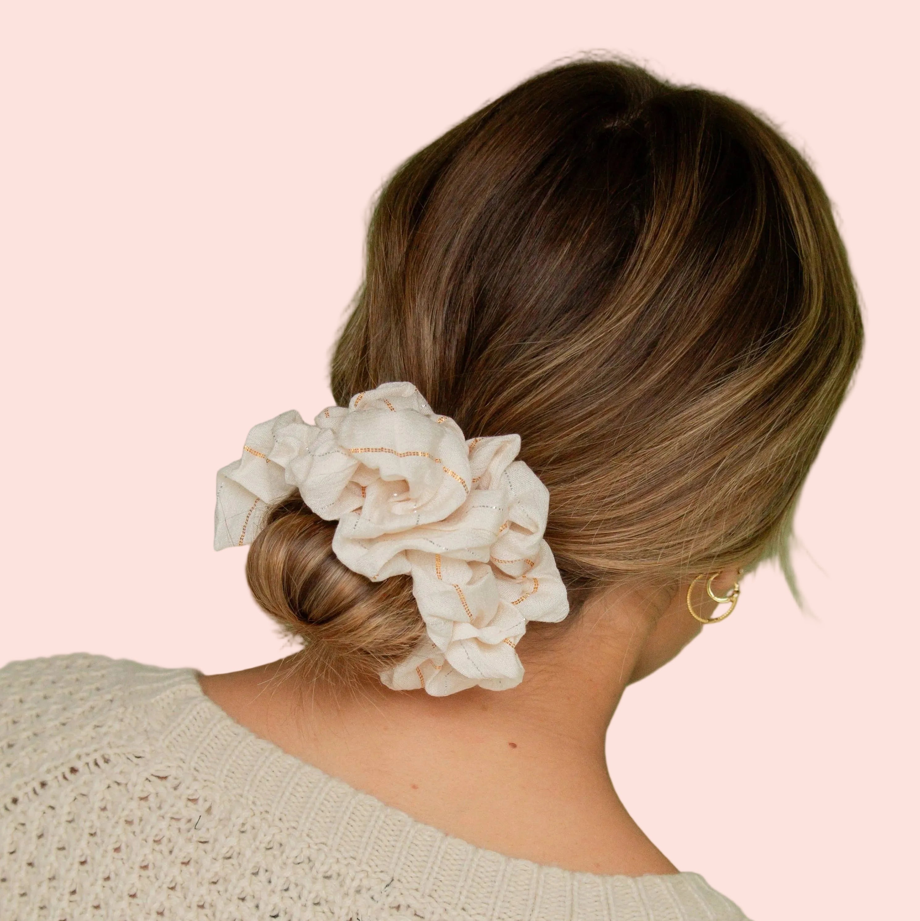 A woman shown from the back wearing a large cream fabric flower hair scrunchie in her brown hair, set against a solid sage green background.