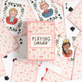 A flat-lay of a pink and white playing card deck box labeled 'PLAYING cards' surrounded by illustrated playing cards on a white background.