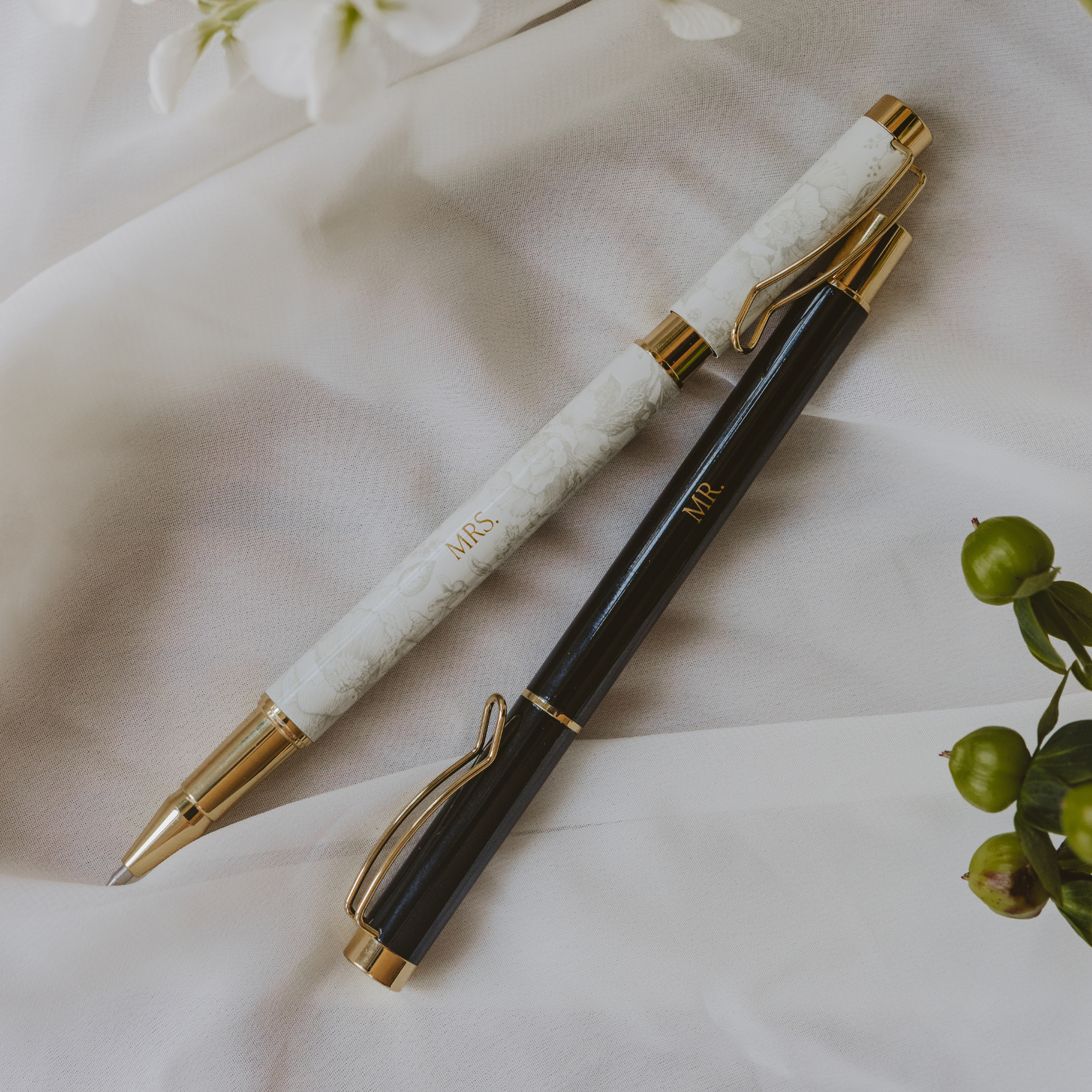 Two pens, one white with gold accents and the other black with gold accents, on a light fabric background.