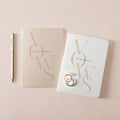 Husband and Wife wedding vow books with gold foil details, matching gold pen, and wedding rings on a soft blush background.