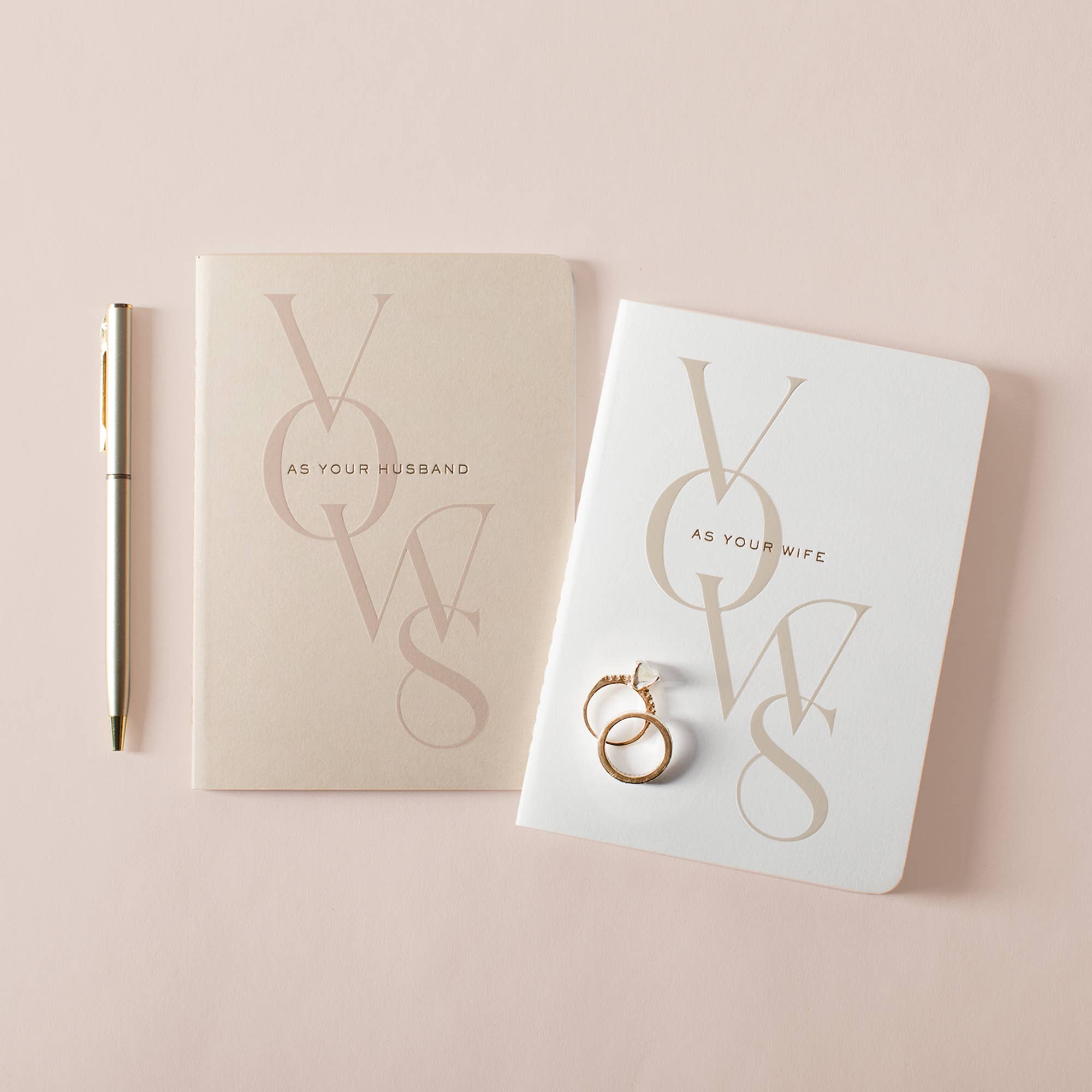 Husband and Wife wedding vow books with gold foil details, matching gold pen, and wedding rings on a soft blush background.