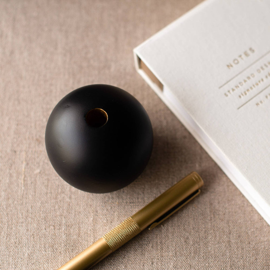 A matte black spherical object with a small hole, styled on a beige textured surface next to a gold pen and a white notebook.