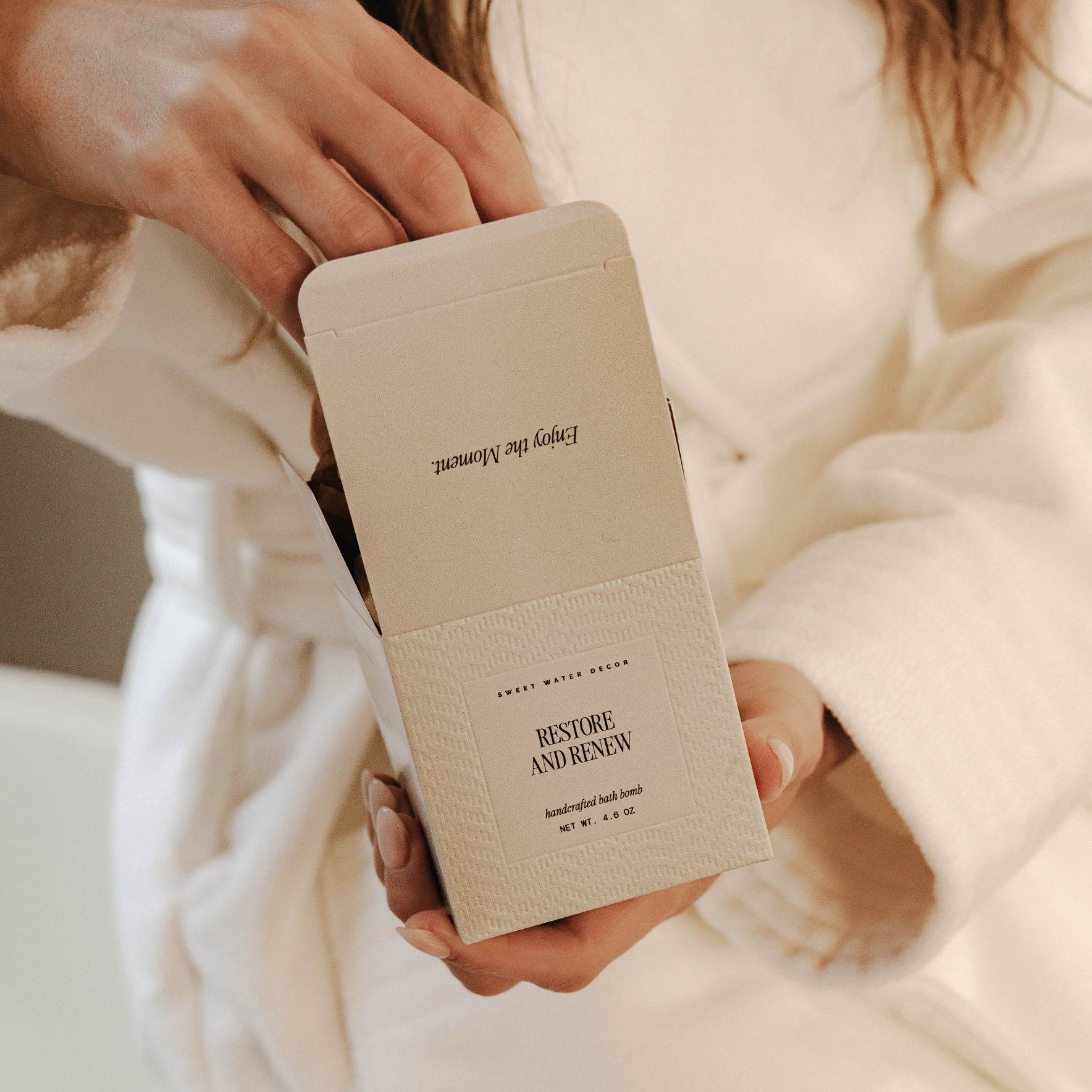 Person holding a beige product packaging with text, wearing a white robe.