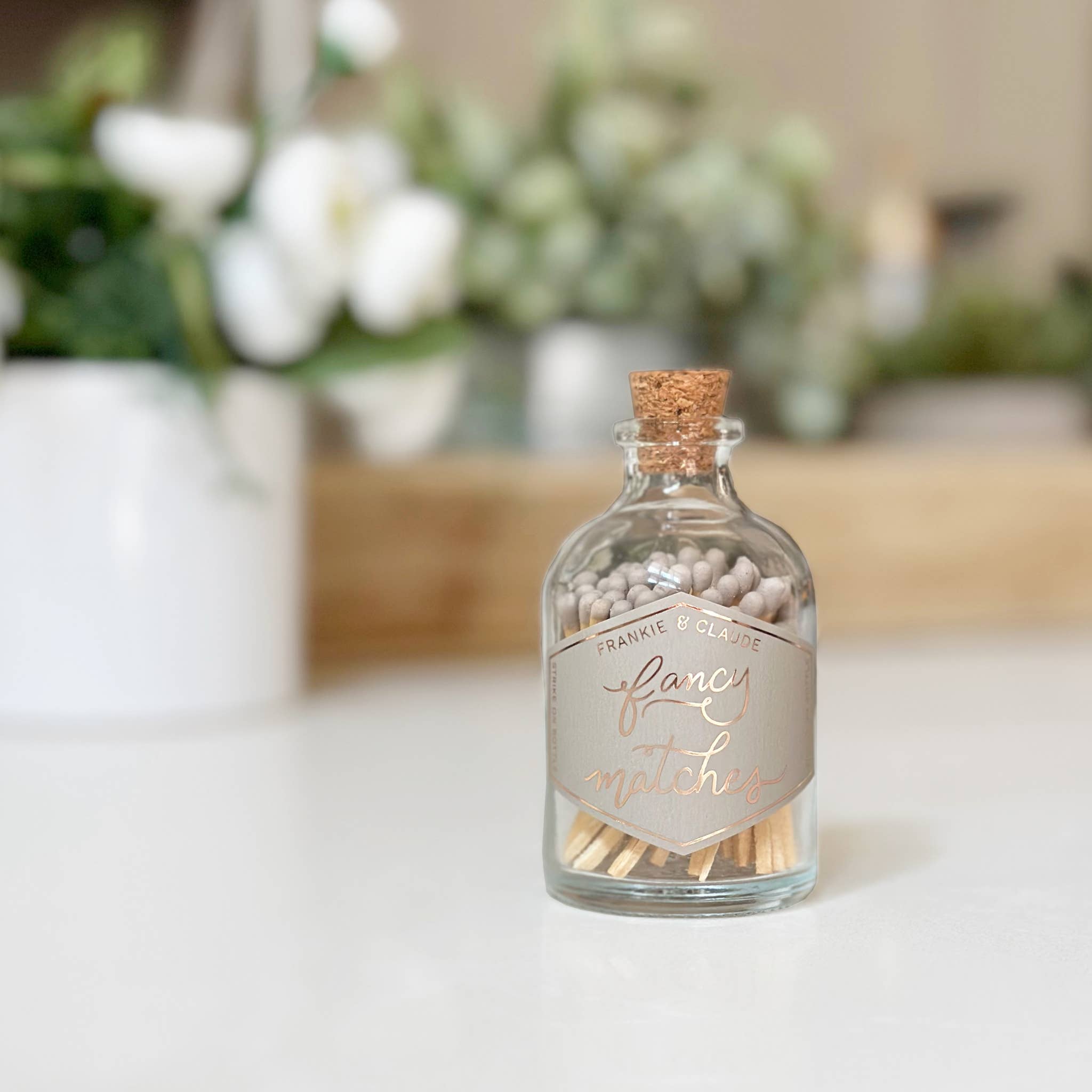 Glass bottle with cork lid containing matches labeled 'fancy' on a white surface with blurred greenery in the background.