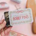 Hand holding a small tin labeled 'emergency bobby pins' with floral design.