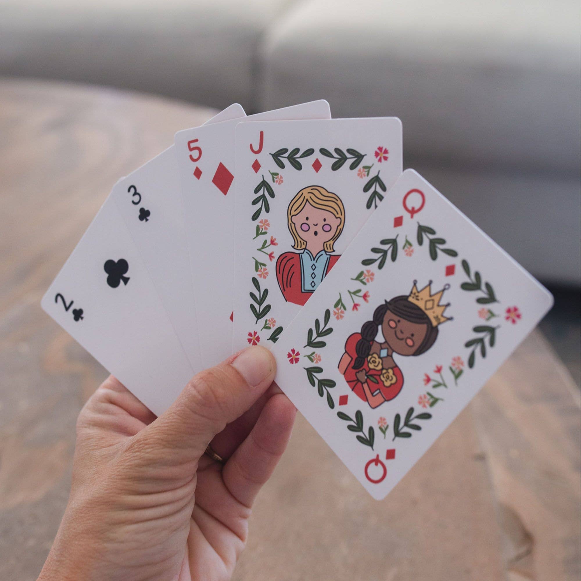 A hand holds four illustrated playing cards fanned out against a blurred indoor background. The cards are a 2 of clubs, 3 of clubs, 5 of diamonds, and a jack of diamonds, featuring cartoon characters and floral leaf borders.