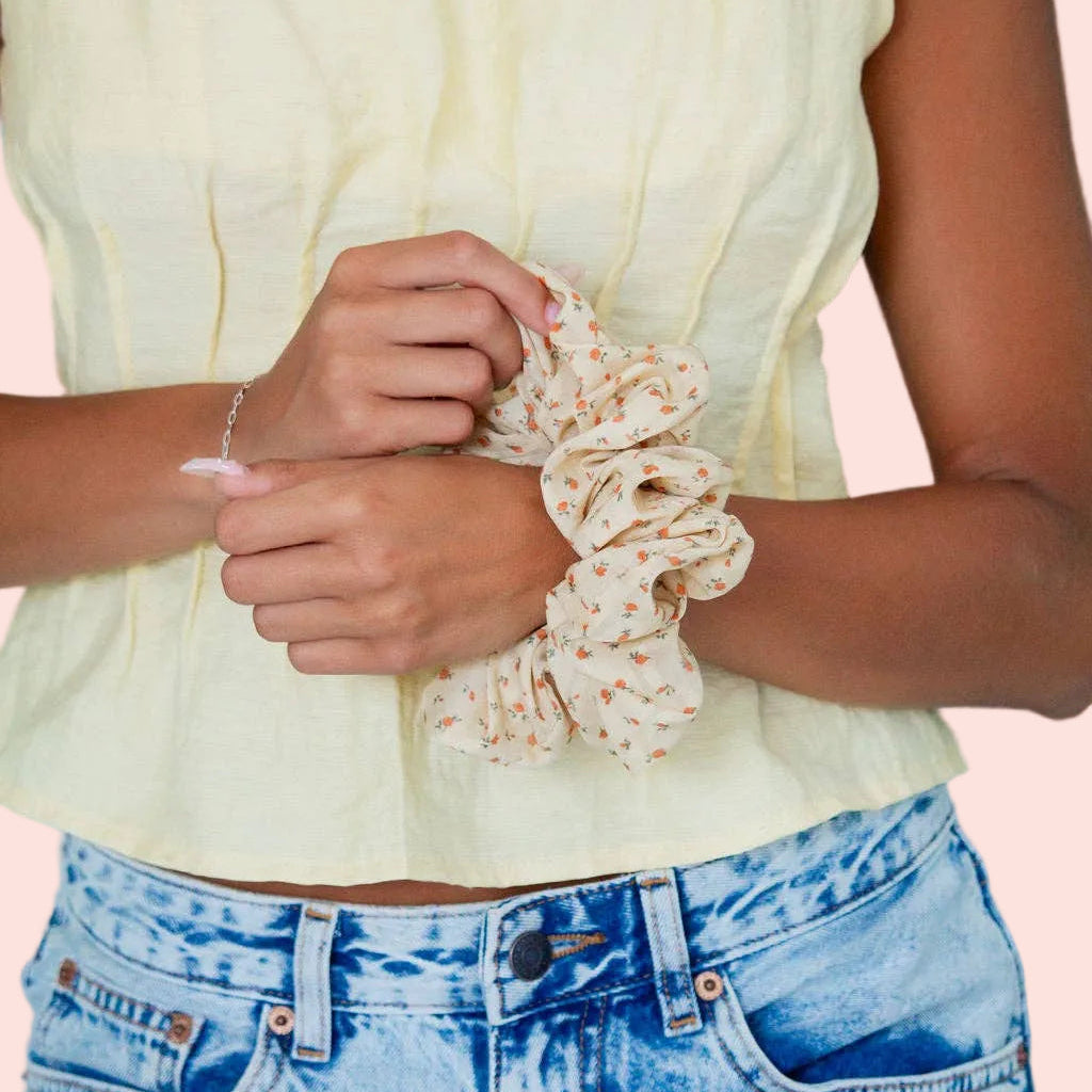 A close-up of a person wearing a pale yellow sleeveless top and light blue jeans, holding a cream scrunchie with a small orange floral pattern against a teal background.