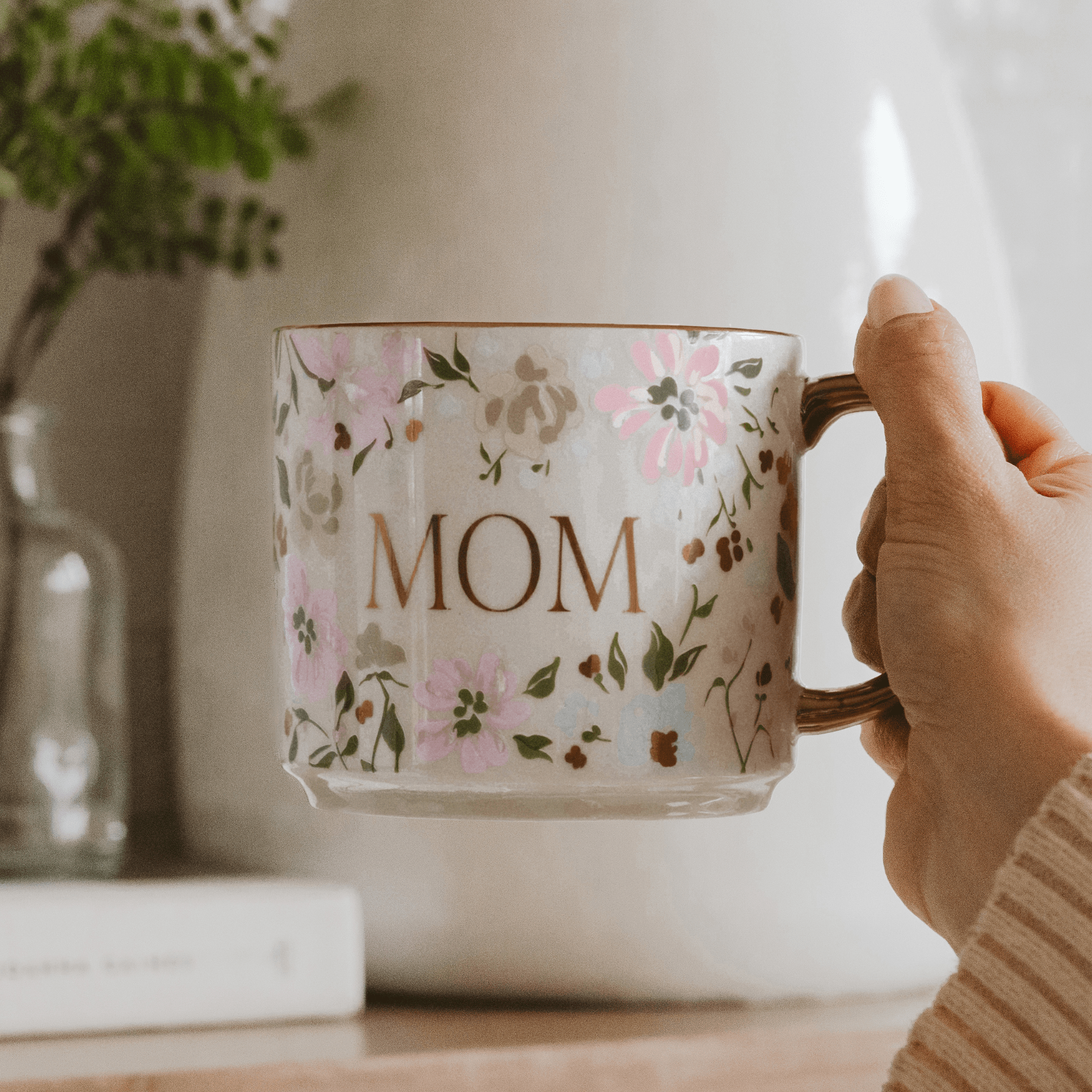 Hand holding a floral mug with 'MOM' printed on it, placed on a wooden surface.