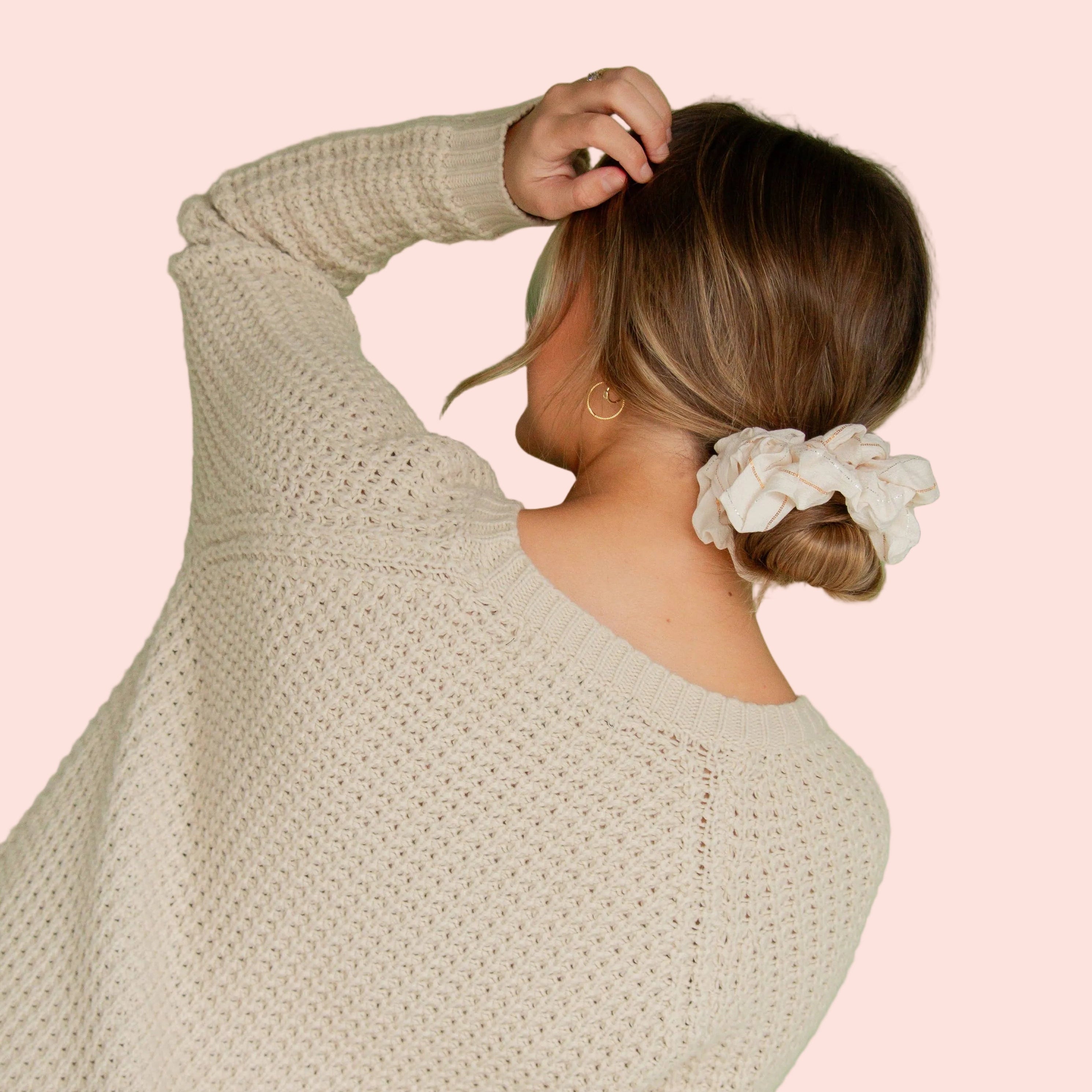 A woman with her hair in a low bun wearing a cream-colored knit sweater and a matching cream fabric scrunchie, shown from behind against a solid sage green background.