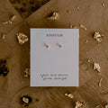 Pair of earrings on a card with text, surrounded by decorative elements on a textured surface