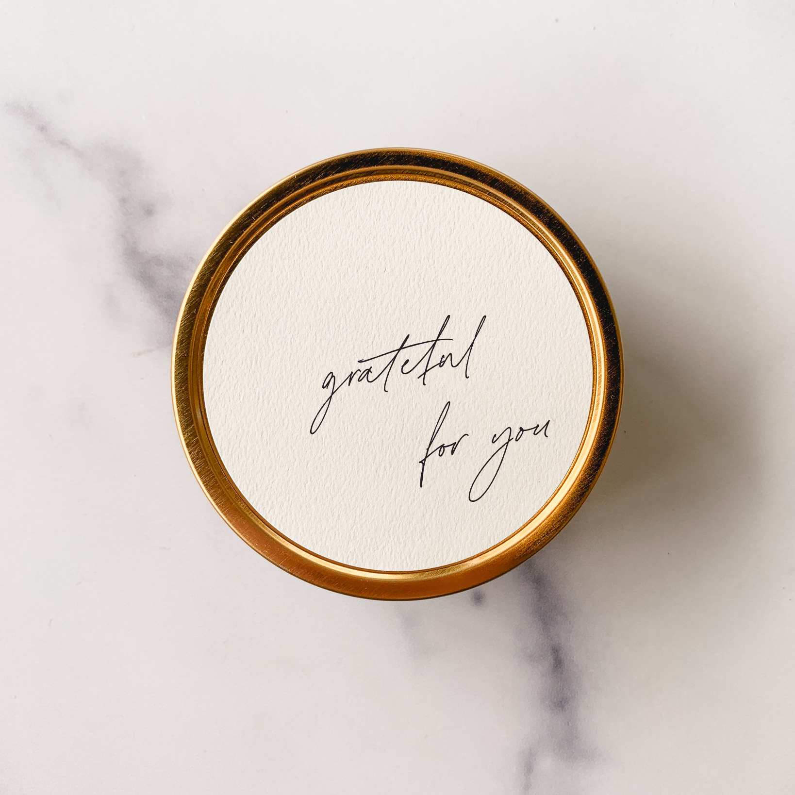 Round white coaster with 'grateful for you' text on a gold rim, placed on a marble surface.