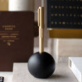 A gold pen with a knurled grip stands upright in a matte black spherical holder on a tabletop, with blurred books and office items in the background.