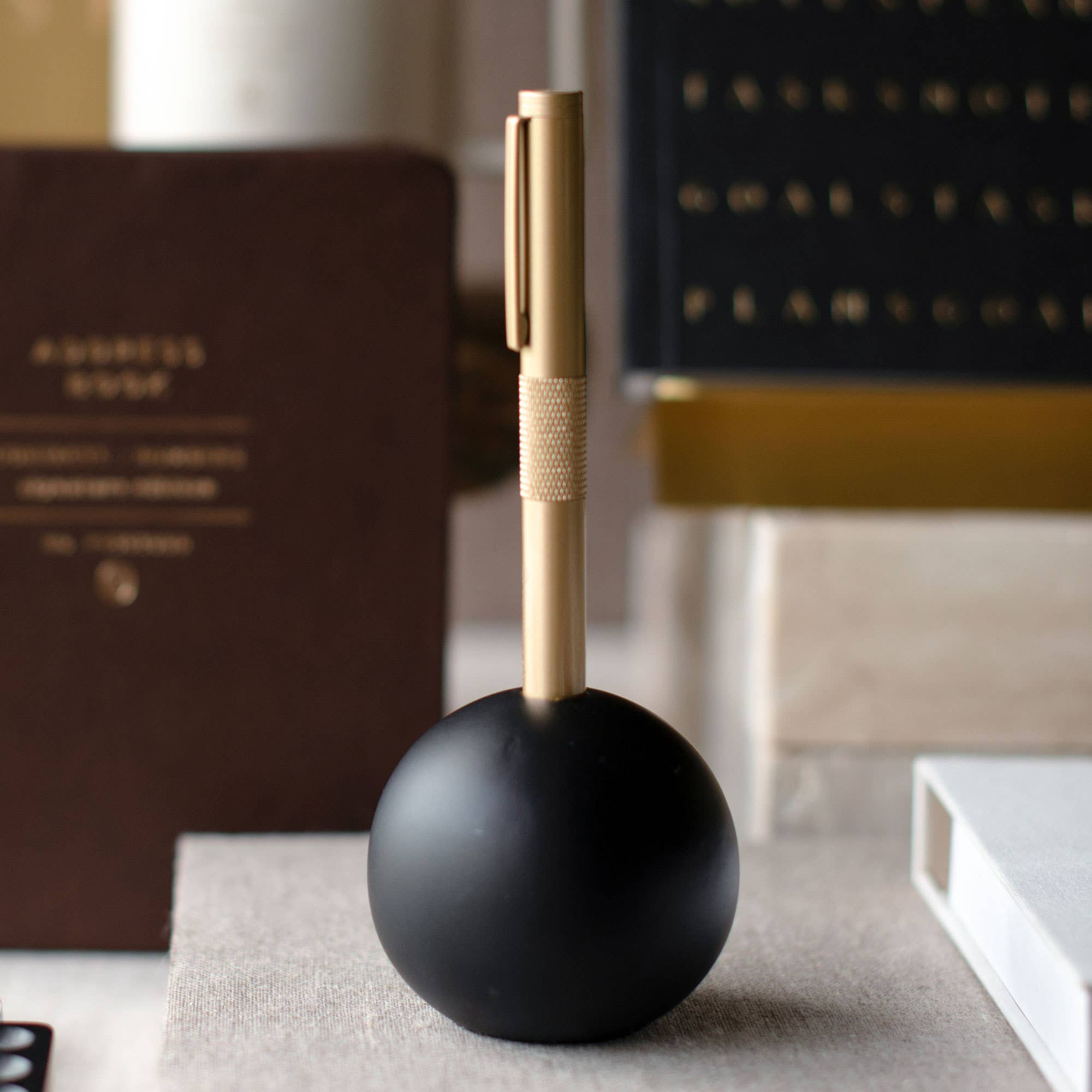 A gold pen with a knurled grip stands upright in a matte black spherical holder on a tabletop, with blurred books and office items in the background.
