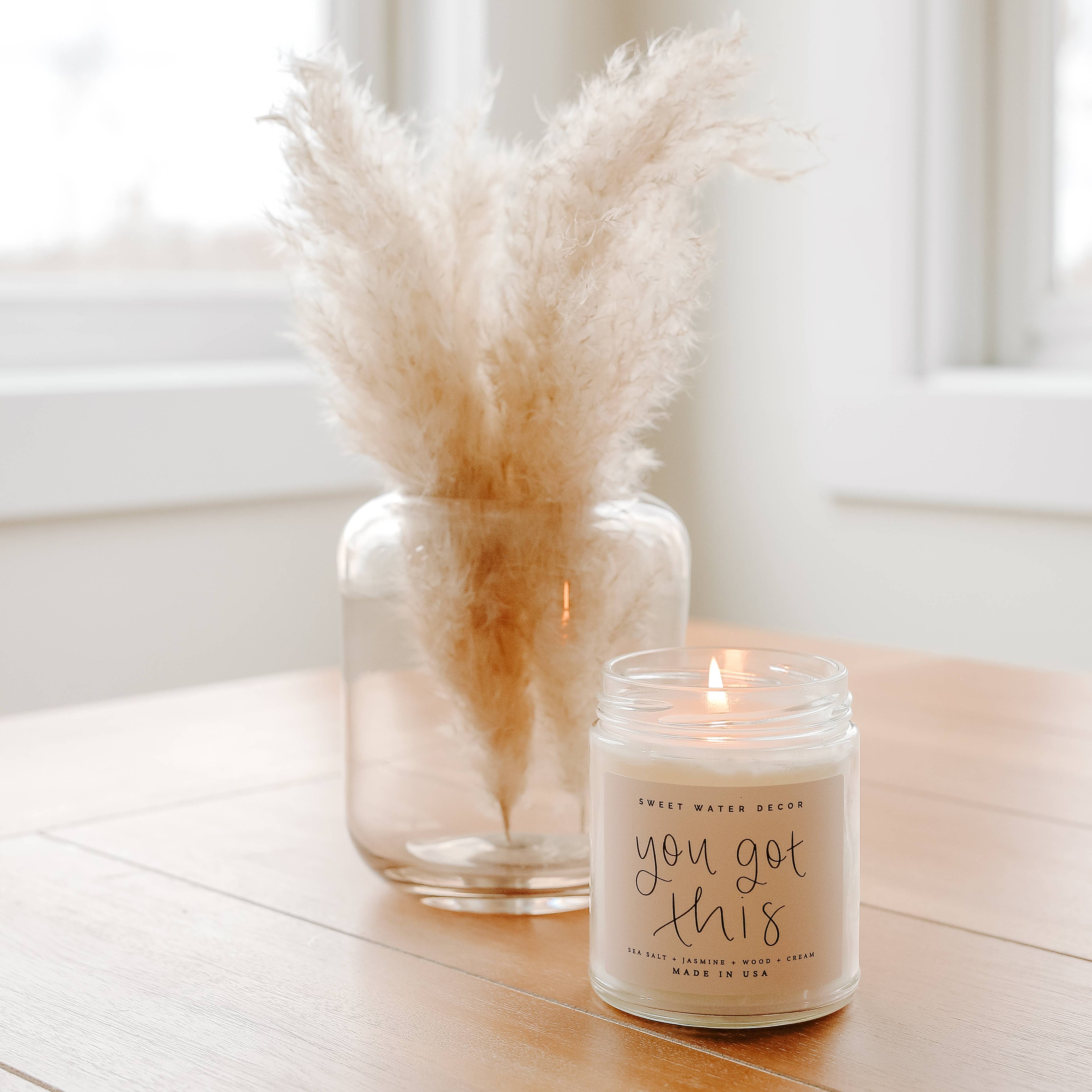 Candle with 'you got this' text and a vase with dried pampas grass on a wooden surface.