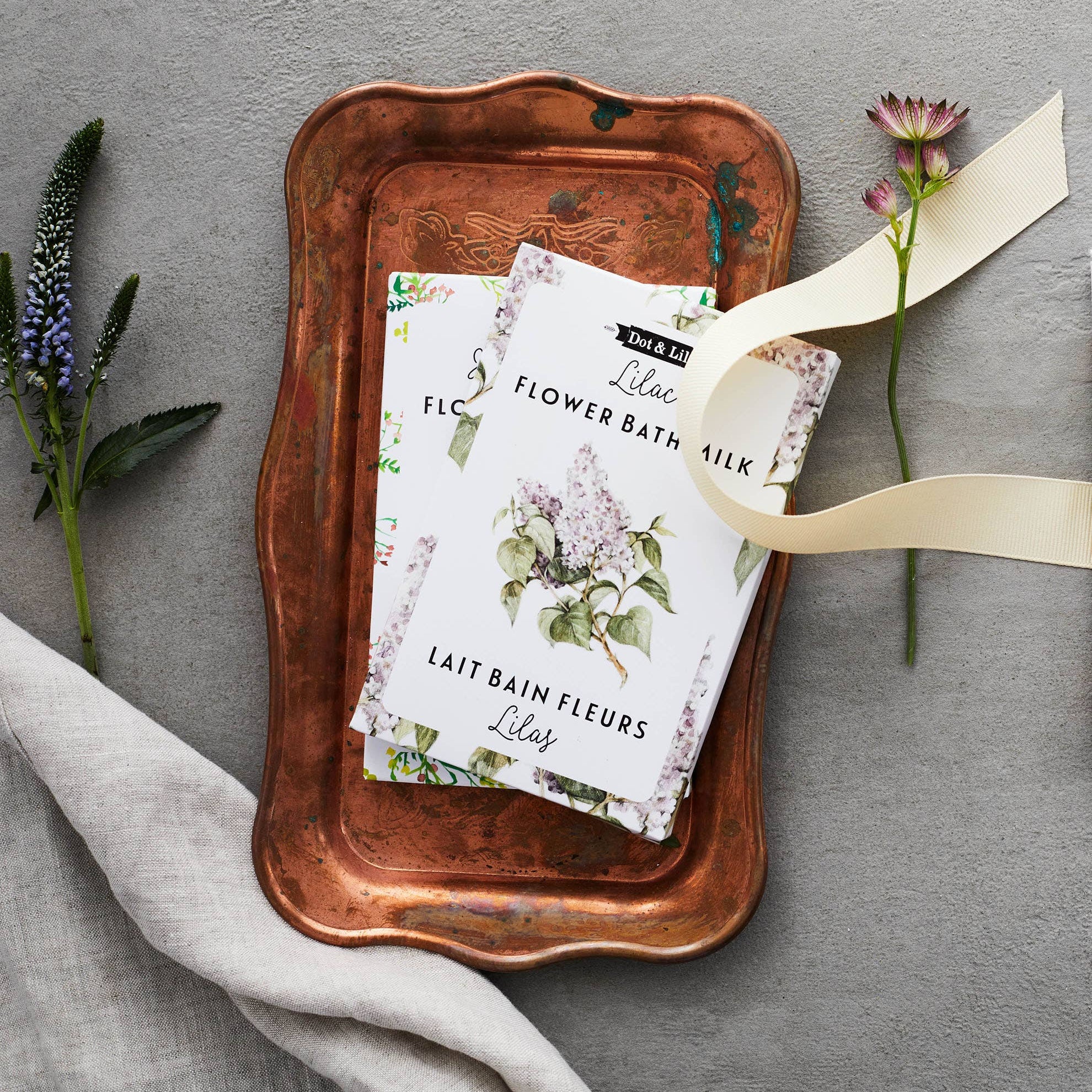 Flower bath milk packaging on a copper tray with flowers and ribbon on a gray background