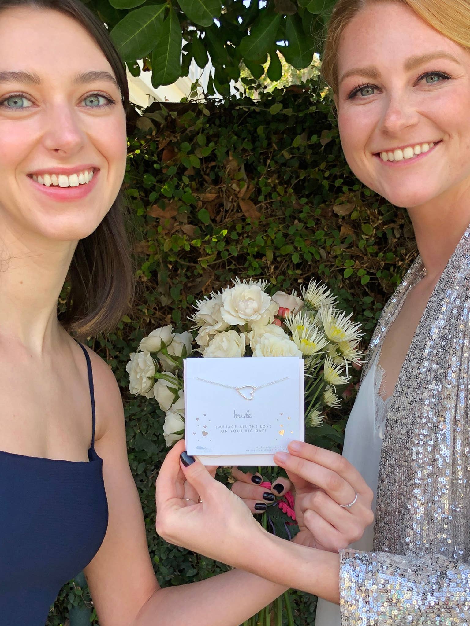 Two women holding a jewelry box with a necklace inside, surrounded by flowers and greenery.