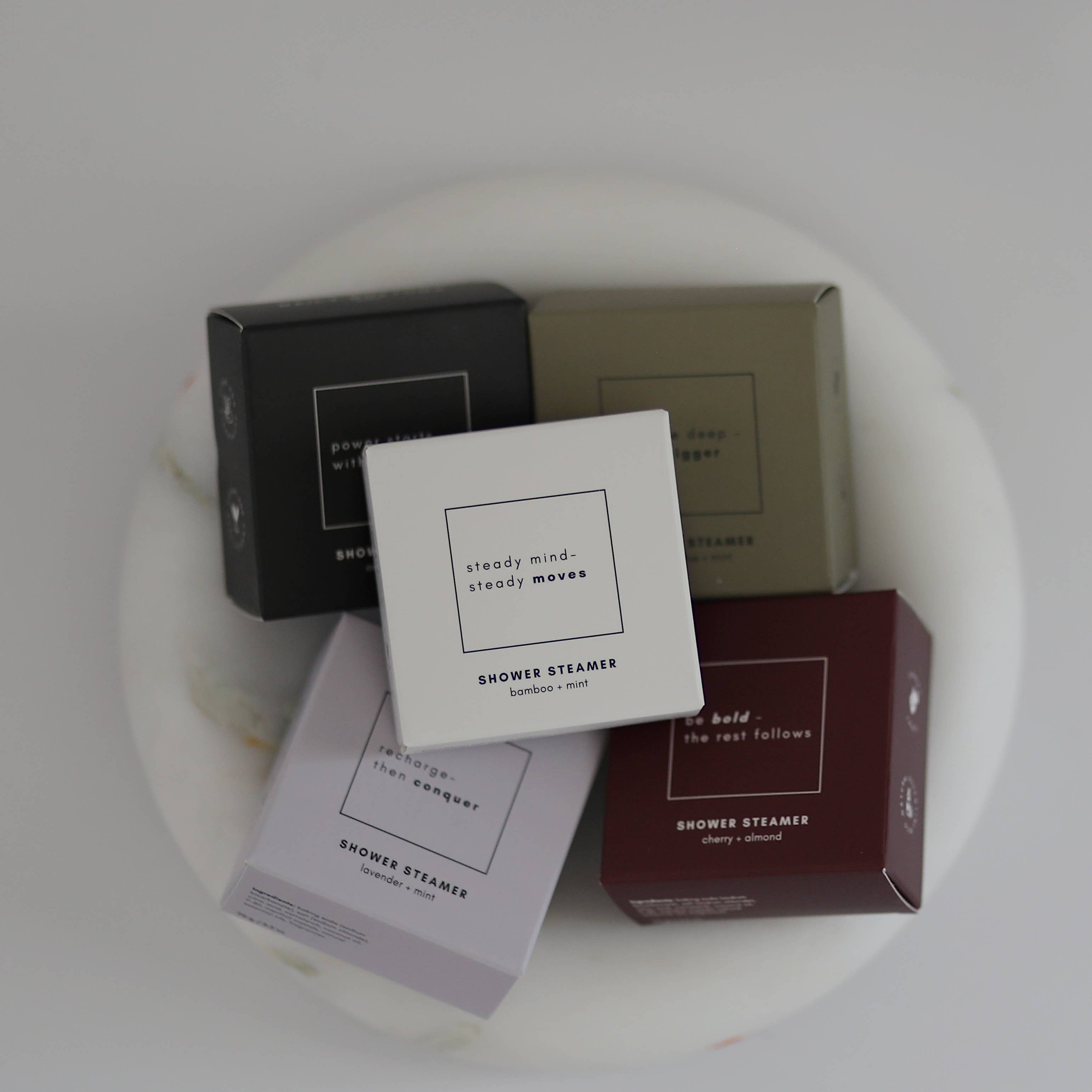 Five small square boxes of SMOOKY STEAMERS body products arranged on a white round tray against a light gray background. The boxes are black, beige, white, pale lavender, and deep burgundy, each with minimalist text on the front.