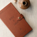 A brown textured leather notebook with a snap-button closure, embossed with the words 'NOTES & IDEAS', photographed on a light linen surface with a small ceramic vase prop.