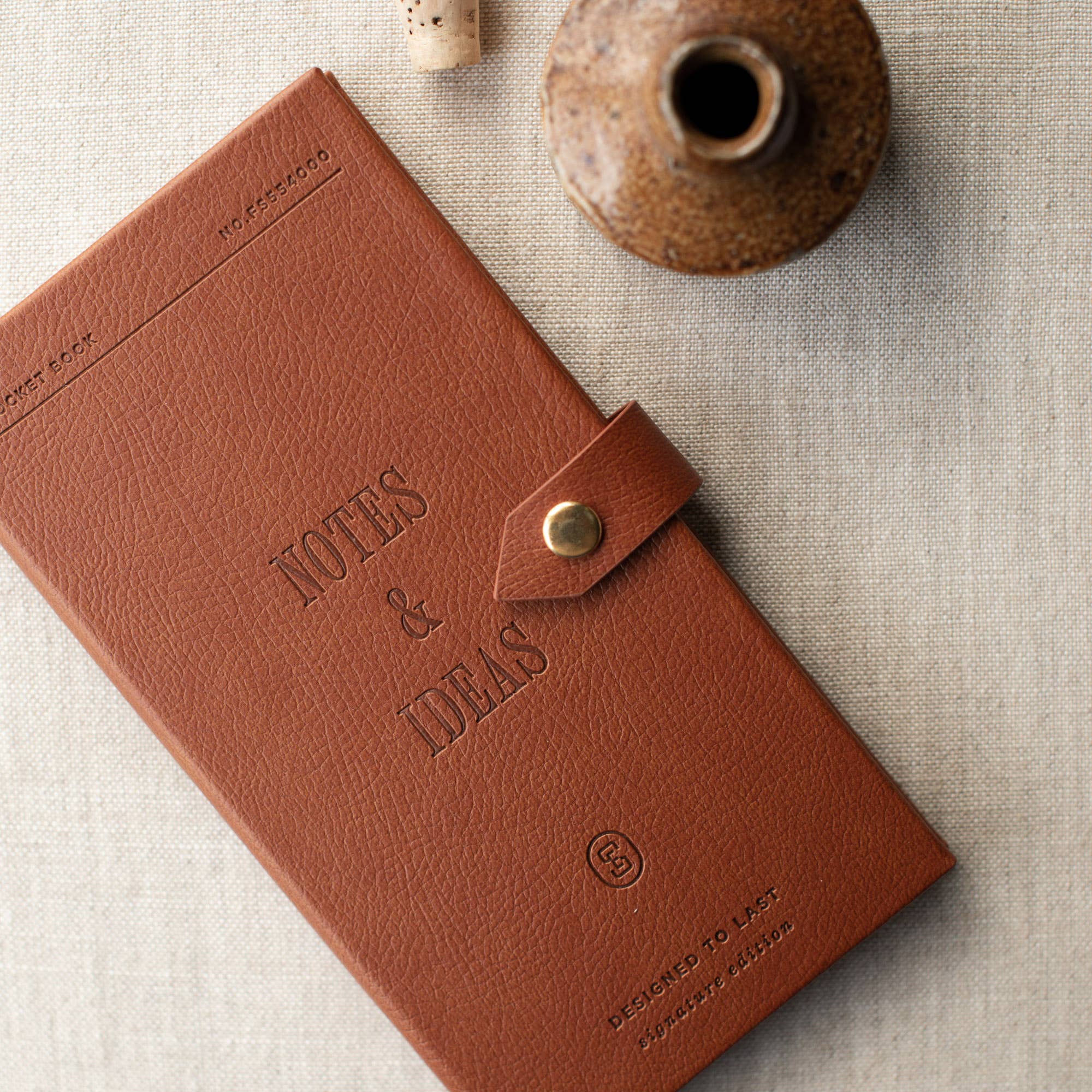 A brown textured leather notebook with a snap-button closure, embossed with the words 'NOTES & IDEAS', photographed on a light linen surface with a small ceramic vase prop.
