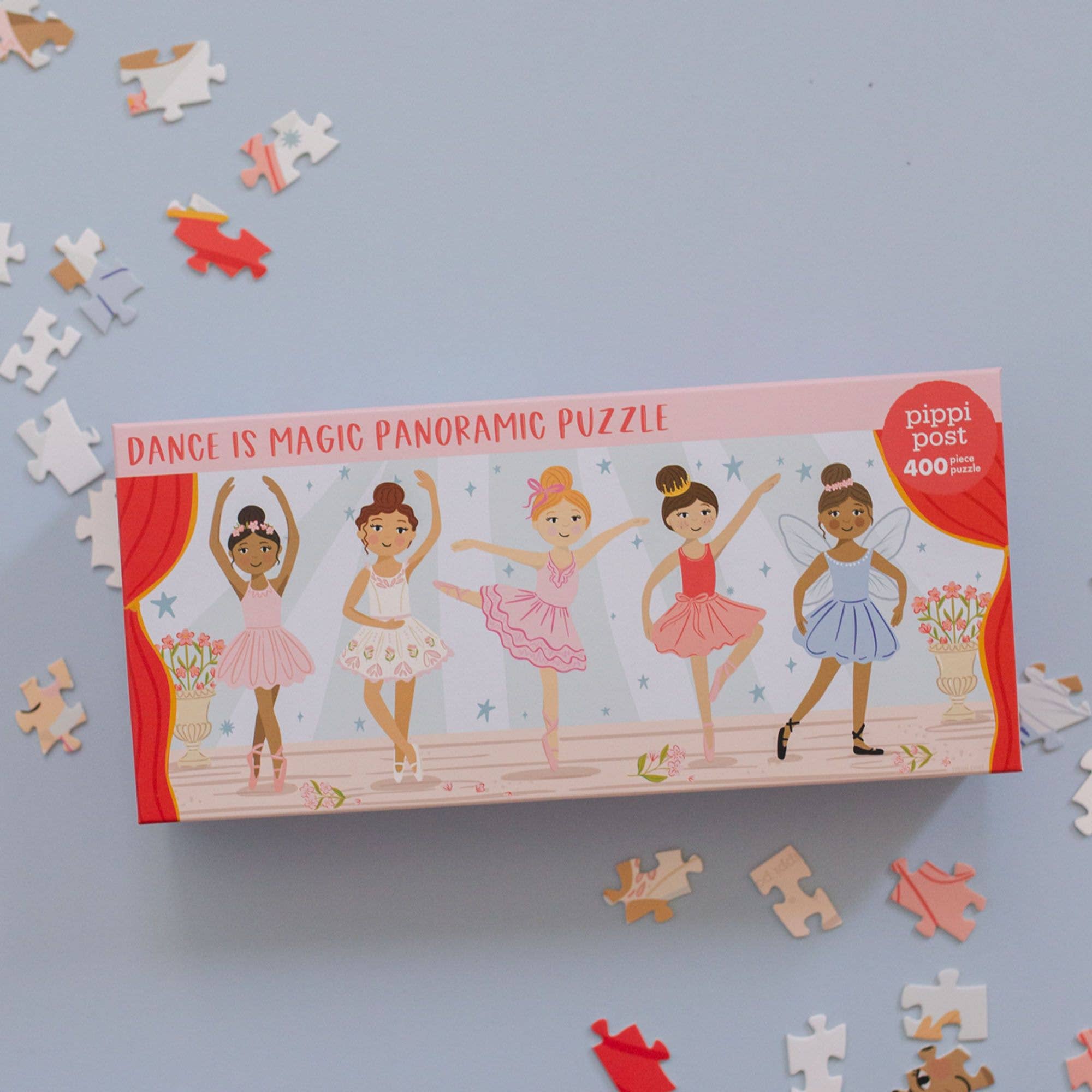 A boxed jigsaw puzzle labeled 'Dance is Magic Panoramic Puzzle' with five ballerina illustrations on the front, photographed on a light blue surface with scattered puzzle pieces around it.