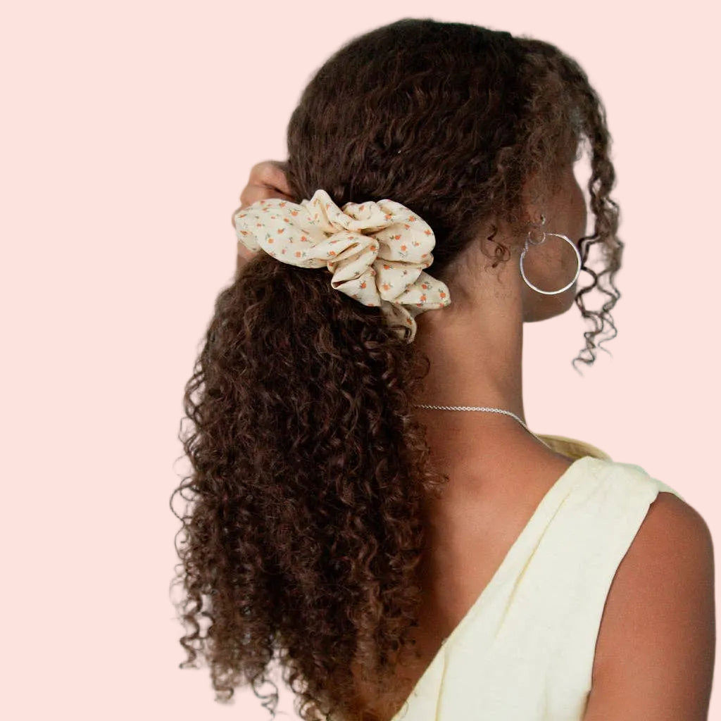 A person shown from the back wearing a cream-colored fabric scrunchie with a small floral pattern, styled with curly brown hair, against a solid light blue background.