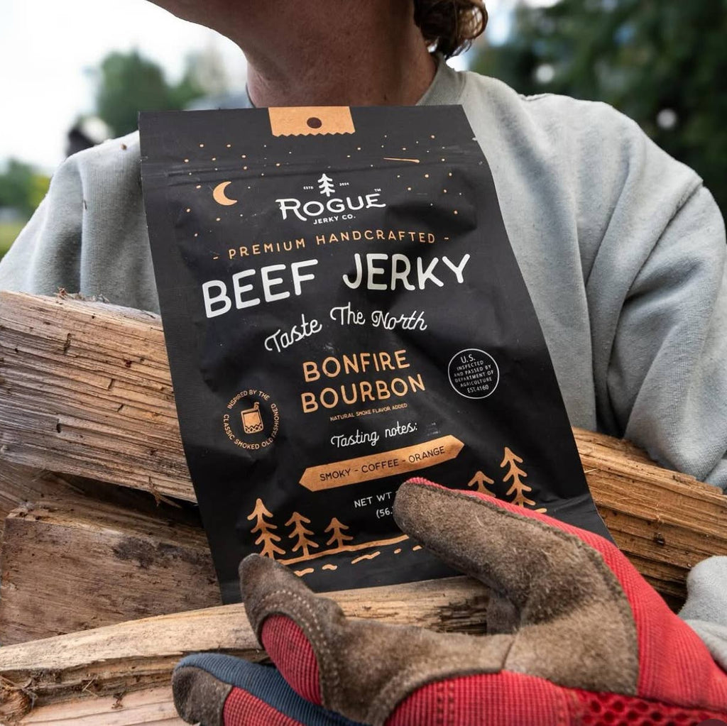 Person holding a package of Rogue Beef Jerky with gloves on, standing next to firewood.