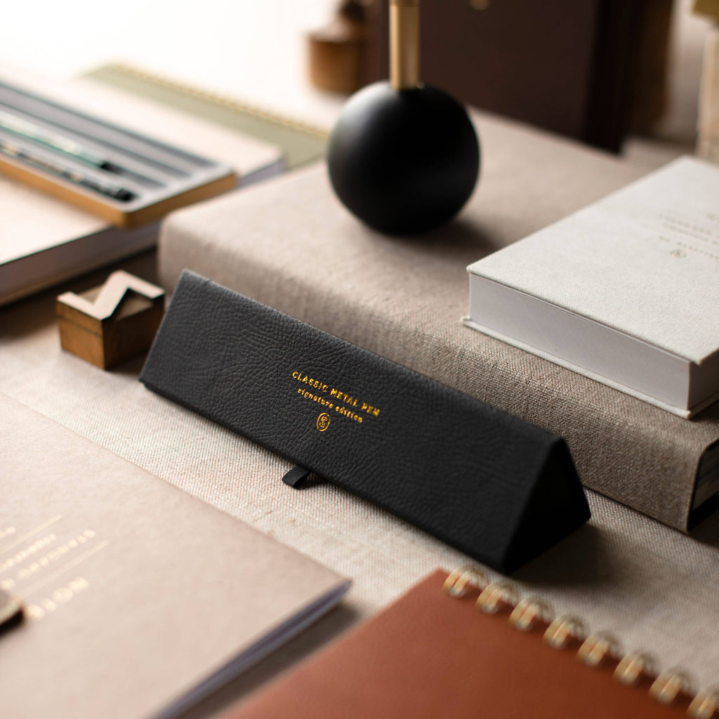A black textured leather eyeglasses case with gold lettering reading 'CLASSIC BOTTLE NO. 05' and a small shield logo, resting on a beige tabletop surrounded by books and stationery.