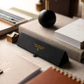 A black textured leather eyeglasses case with gold lettering reading 'CLASSIC BOTTLE NO. 05' and a small shield logo, resting on a beige tabletop surrounded by books and stationery.