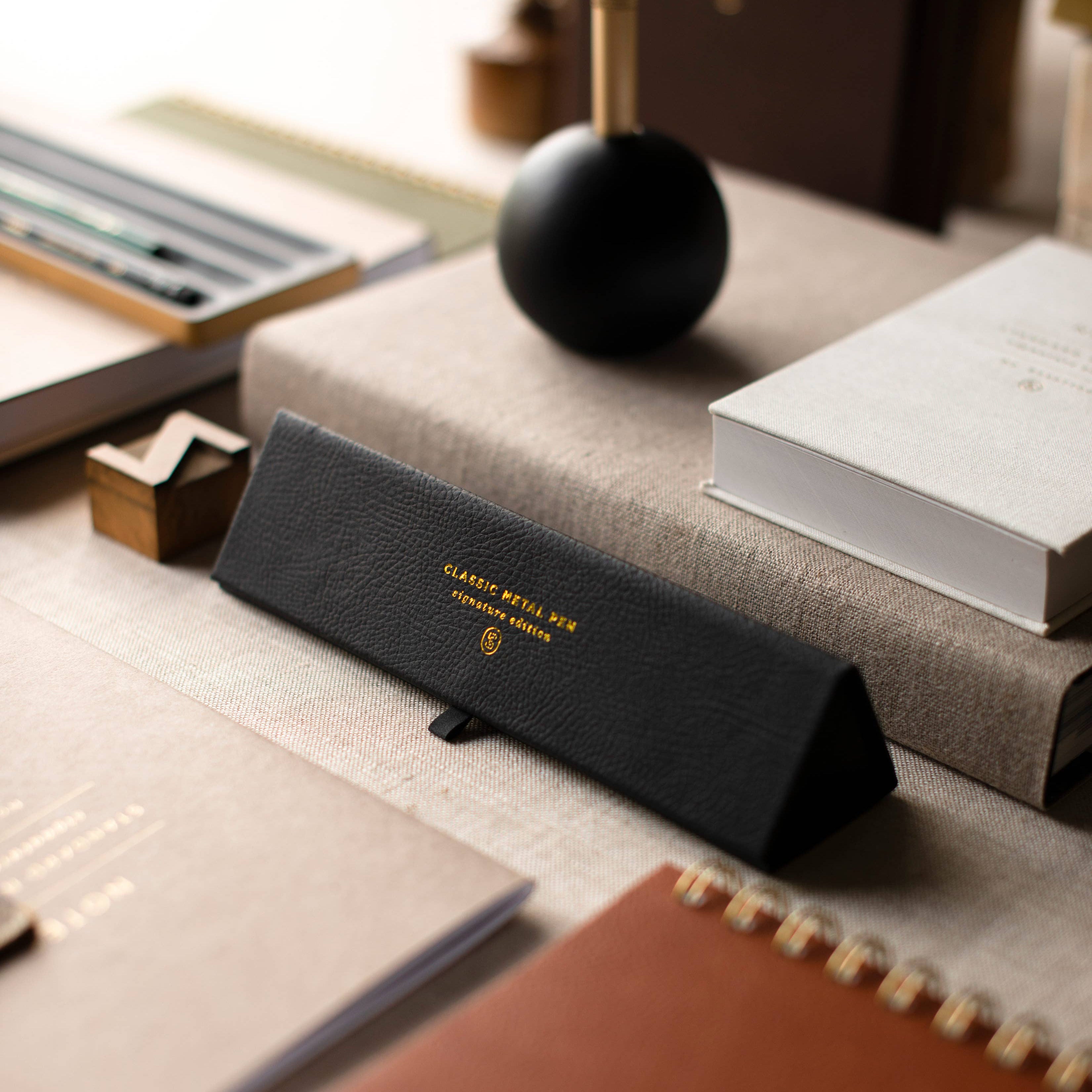 A black textured leather eyeglasses case with gold lettering reading 'CLASSIC BOTTLE NO. 05' and a small shield logo, resting on a beige tabletop surrounded by books and stationery.