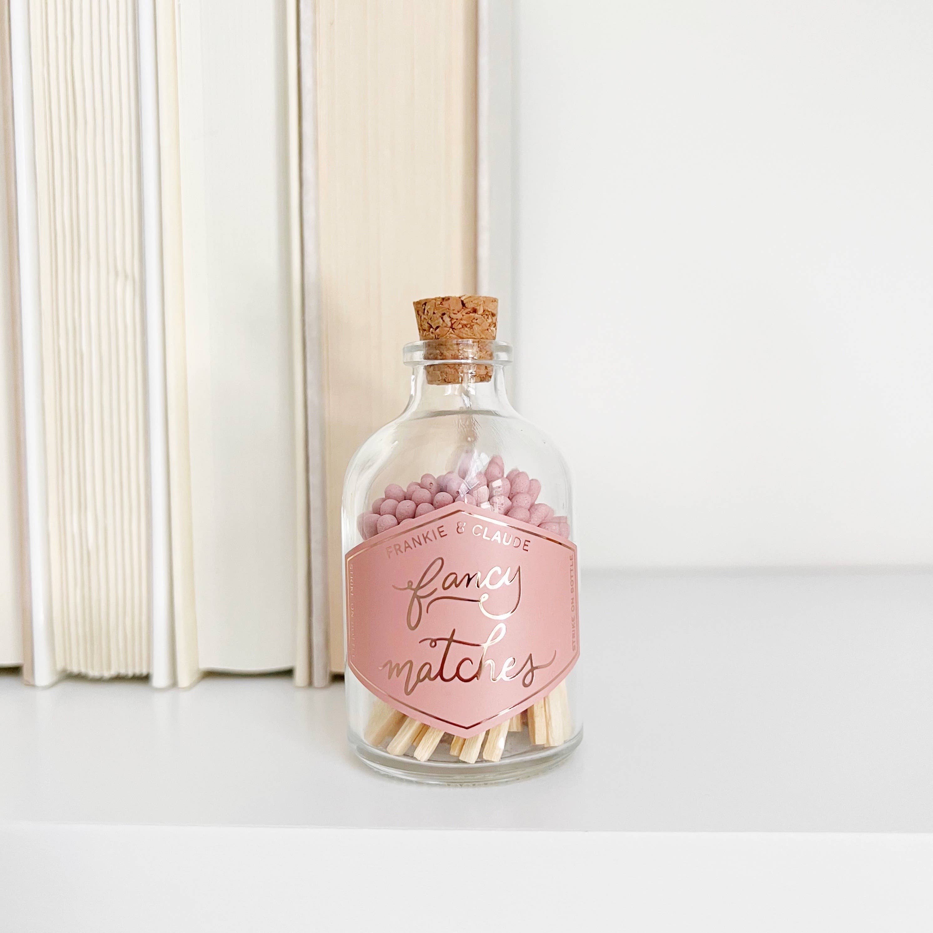 Glass bottle with cork lid containing pink matches and a label on a white surface.