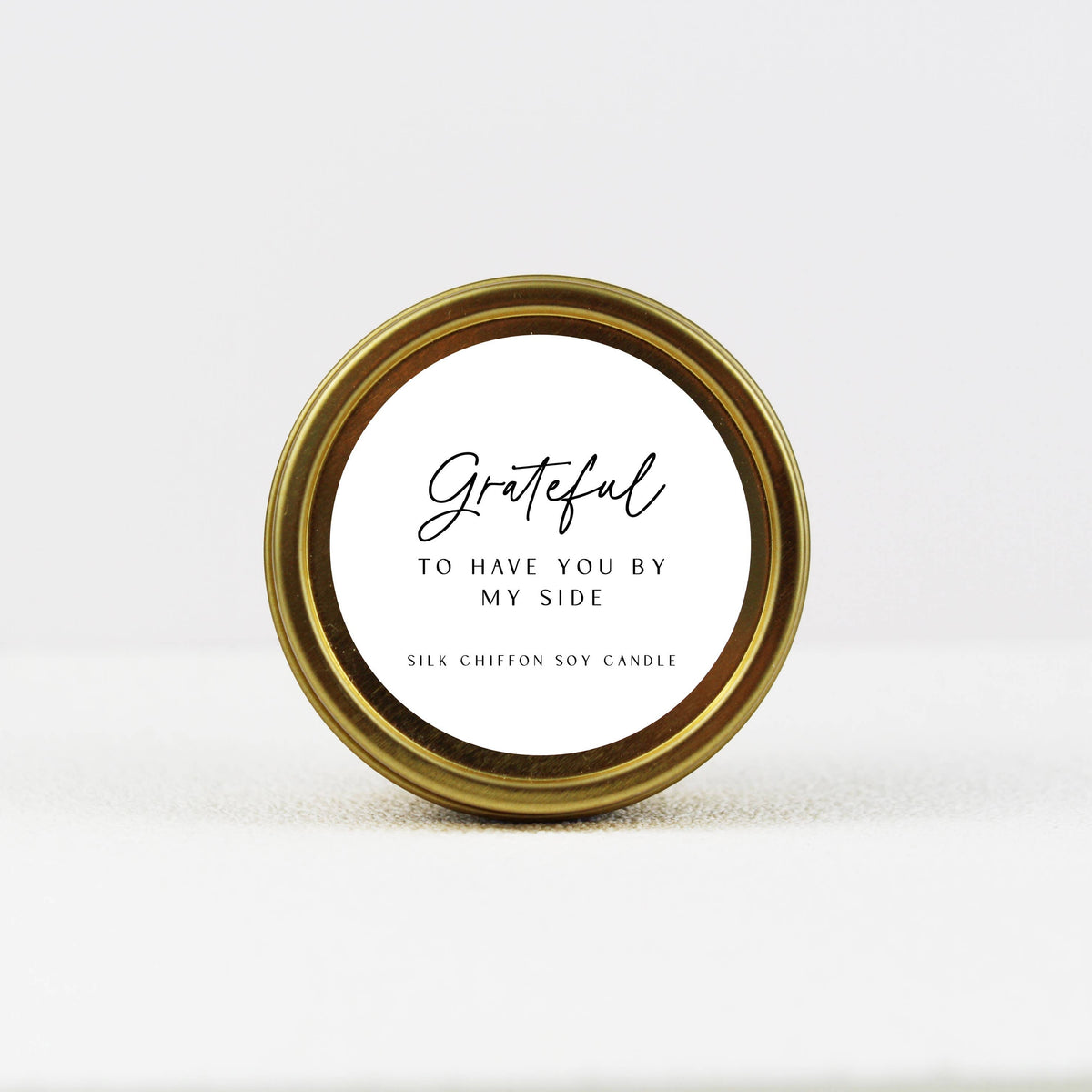 A round gold metal tin candle lid with a white paper label that reads 'Grateful TO HAVE YOU BY MY SIDE' and 'SILK CHIFFON SOY CANDLE' in black text, photographed on a clean white background.