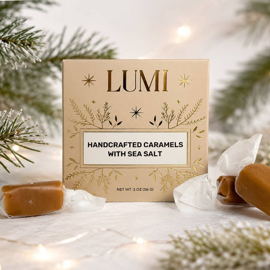 LUMI Handcrafted Caramels with Sea Salt in a beige box with gold leaf details, styled with unwrapped caramel candies and holiday pine branches on a snowy white surface with warm string lights.